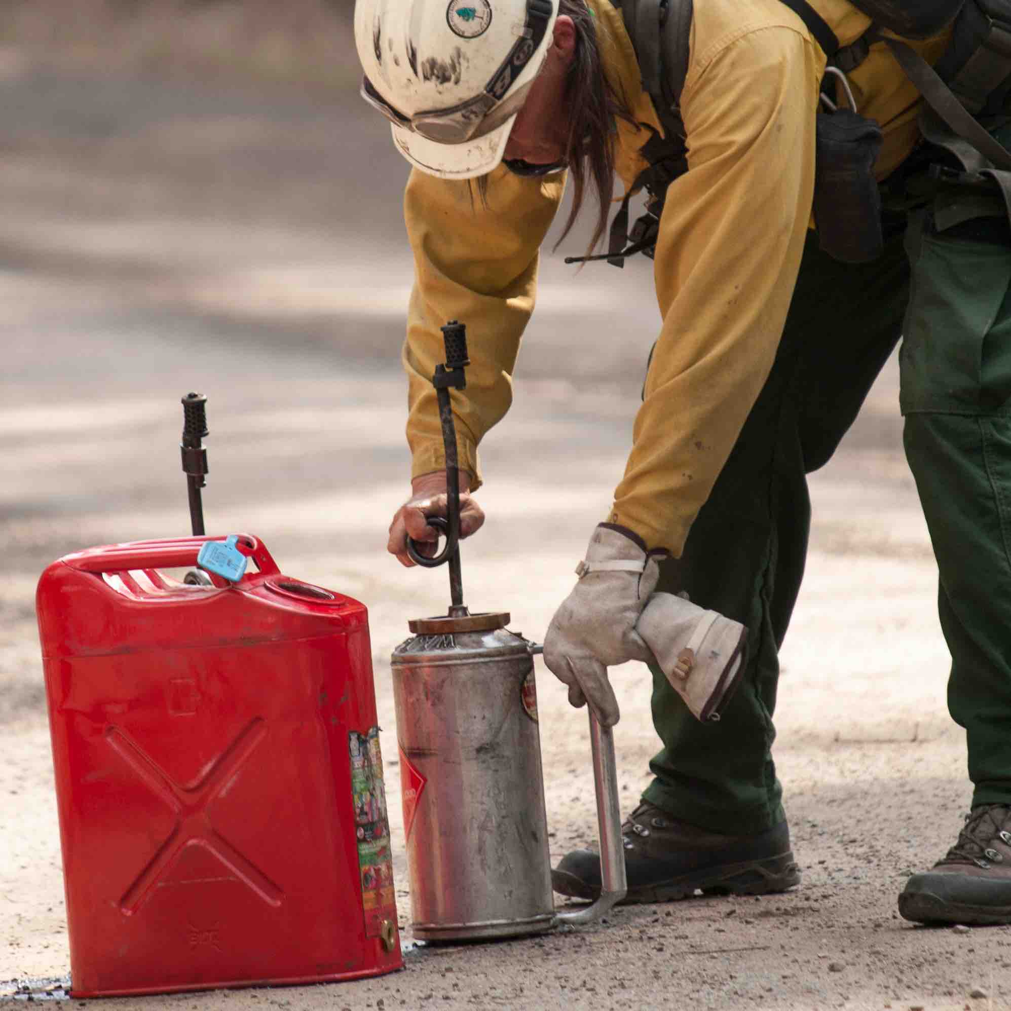 firefighter in helmet and yellows unscrewing a drip torch to refill from nearby gas can.